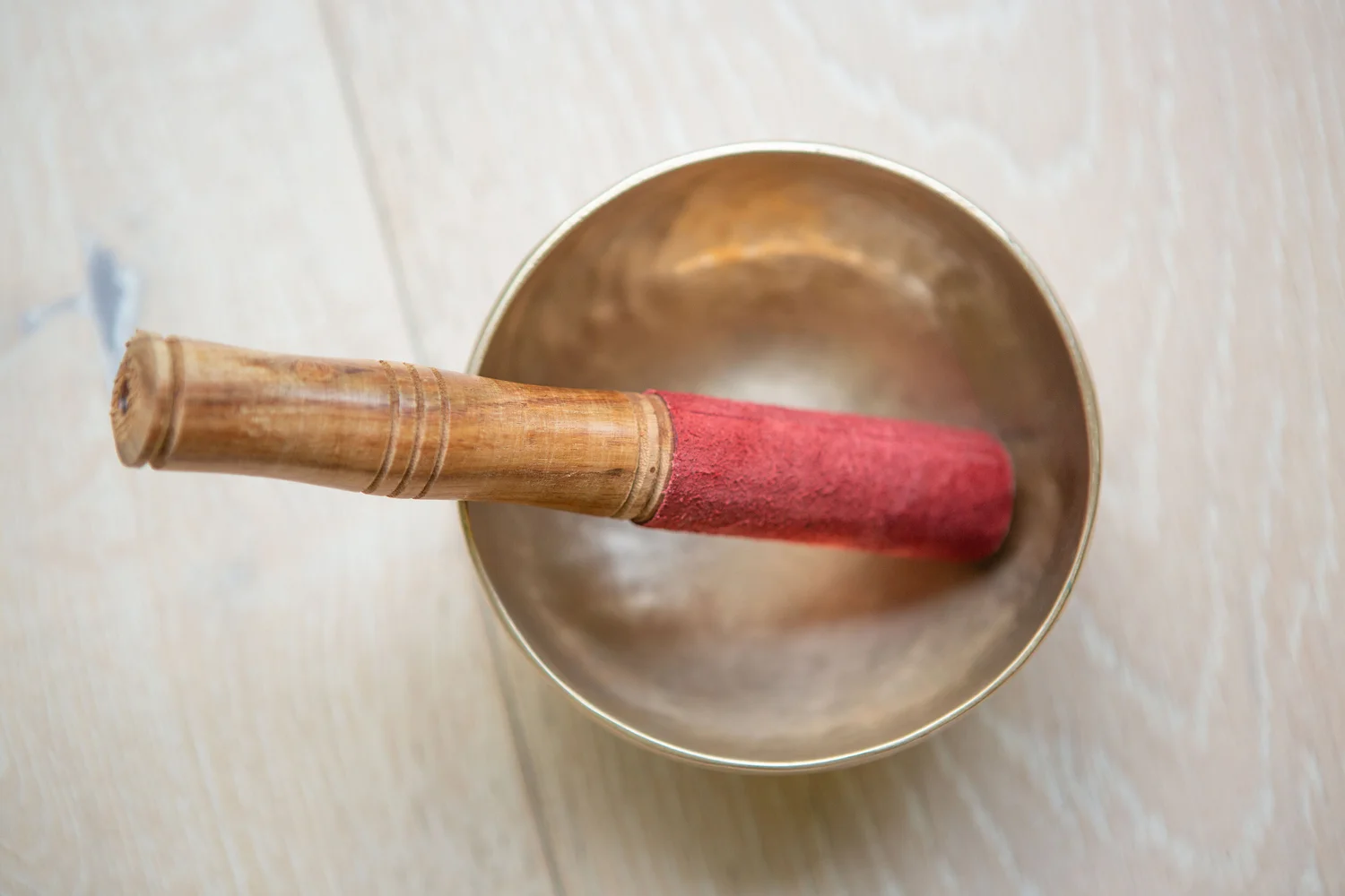 Top-down close-up of a brass singing bowl resting on a pale wooden floor. A wooden mallet with a red felt-covered tip lies across the rim of the bowl, the handle showing carved rings. The image is softly lit and focused on the bowl and mallet, evoking a meditative, yoga studio vibe.