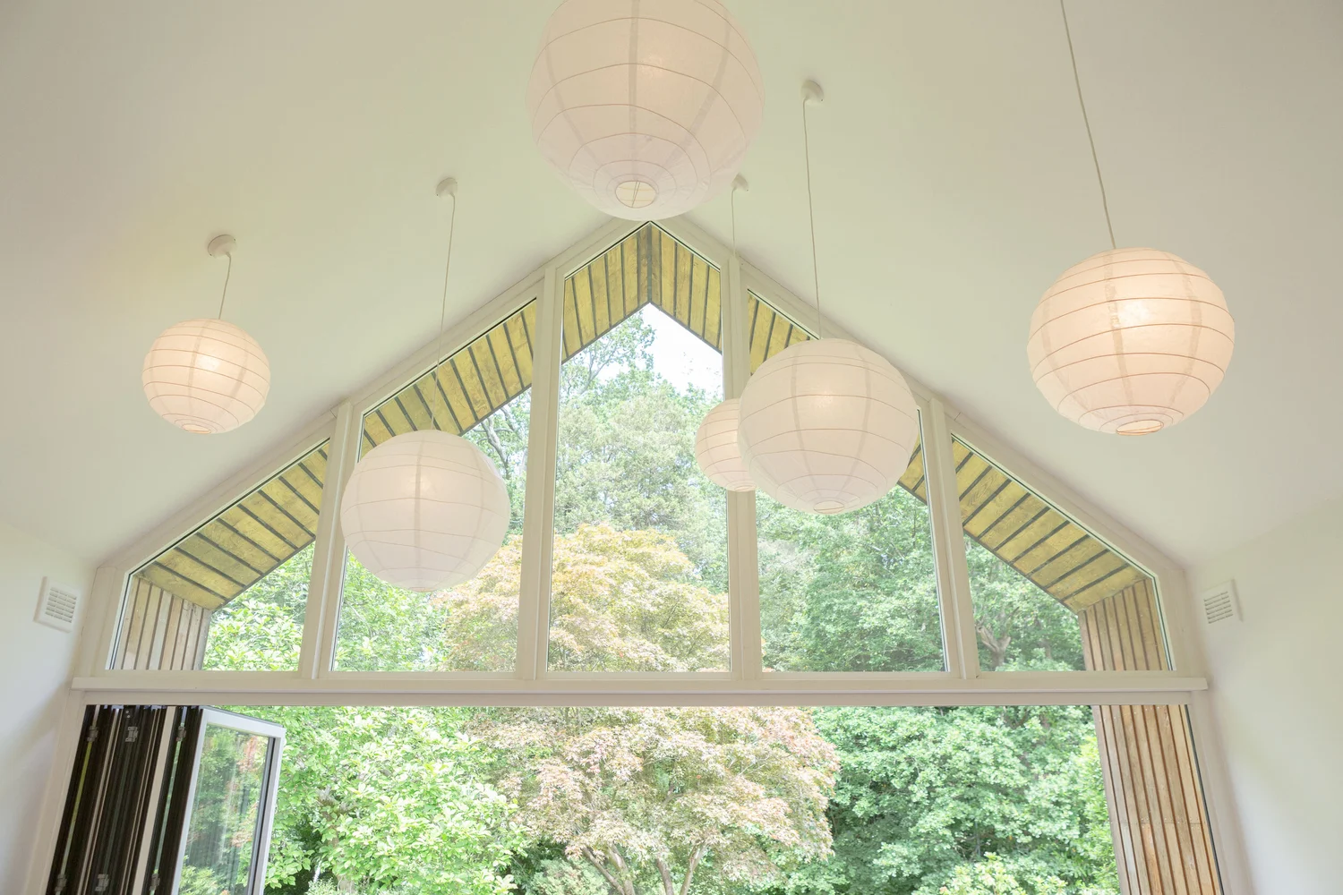 Interior view aimed toward the large gable window of the studio, with green trees visible outside. Several white paper lanterns hang from the high ceiling at different heights, floating in front of the triangular window panes and framing the garden view. The photo emphasizes the airy height, the geometric window shapes, and the soft lantern light.